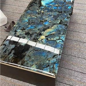Könnte beinhalten: Rechteckiger Couchtisch mit polierter, schillernder Oberfläche. Die Tischplatte weist ein auffälliges Muster aus Blau-, Grün- und Schwarztönen auf, das einem Naturstein ähnelt. Der Tisch hat einen dunkelbraunen Rahmen.