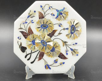 May include: White octagonal marble decorative piece with inlaid floral design. The flowers feature yellow, blue, and brown accents. The piece is supported by a clear stand and is from The Minimalistic Studio.