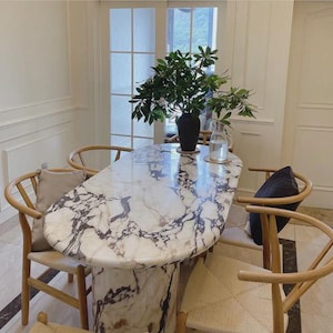 Op de afbeelding: Een ovale marmeren eettafel met een wit en grijs geaderd patroon. De tafel wordt ondersteund door een cilindrische voet. Vier houten stoelen met gebogen rugleuningen omringen de tafel. Een zwarte vaas met groen blad staat op de tafel.