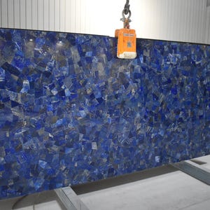 May include: Large slab of blue stone with a mosaic pattern. The stone is a deep blue color with lighter blue and gray accents. It is supported by a metal stand and is being lifted by a crane.