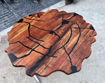 May include: A round live edge wood table top featuring a natural brown grain pattern with irregular edges. The surface is inlaid with black resin filling the cracks and voids in the wood, creating a smooth, continuous, and flat tabletop finish.