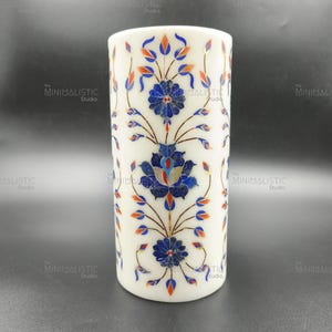 Puede incluir: Un jarrón cilíndrico de mármol blanco con incrustaciones florales. El diseño presenta detalles en azul, naranja y rojo, creando un patrón decorativo. Un artículo de decoración del hogar único.