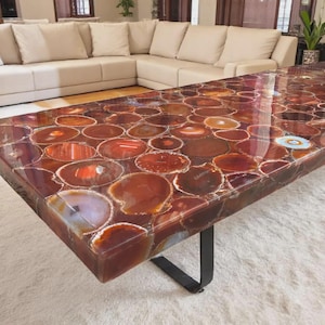 May include: A rectangular coffee table with a polished surface made of agate slices in shades of red, brown, and cream. The table has a black metal base and sits on a white rug. The agate slices are arranged in a mosaic pattern.