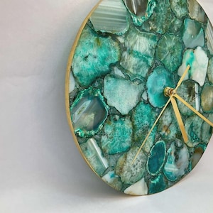 May include: A round wall clock with a natural stone face. The clock face is made of irregularly shaped, polished green and white agate stones. The clock has gold-colored hands and a wooden frame.