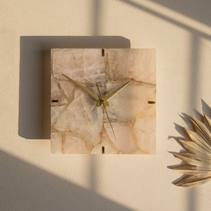 Op de afbeelding: Vierkante wandklok met een lichtroze en beige stenen wijzerplaat. De klok heeft goudkleurige wijzers en kleine markeringen. De klok is bevestigd aan een lichtbeige muur. De wijzerplaat van de klok heeft een natuurlijk, getextureerd uiterlijk.