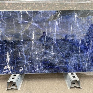 May include: A large rectangular slab of blue marble with white veining. The polished surface reflects light, creating a dynamic pattern. The stone is supported by metal stands, suggesting it's ready for installation as a countertop or wall panel.