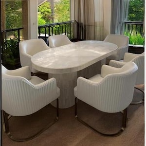 Op de afbeelding: Een moderne eethoek met een witte ovale tafel en zes wit gestoffeerde stoelen met goudkleurige metalen poten. De kamer heeft grote ramen en een kroonluchter.
