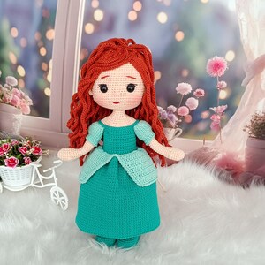 Op de afbeelding: Een handgemaakte gehaakte pop met rood haar en een turquoise jurk. De pop heeft een gedetailleerd gezicht met zwarte ogen en een kleine rode mond. De jurk heeft een gelaagde rok en korte mouwen.