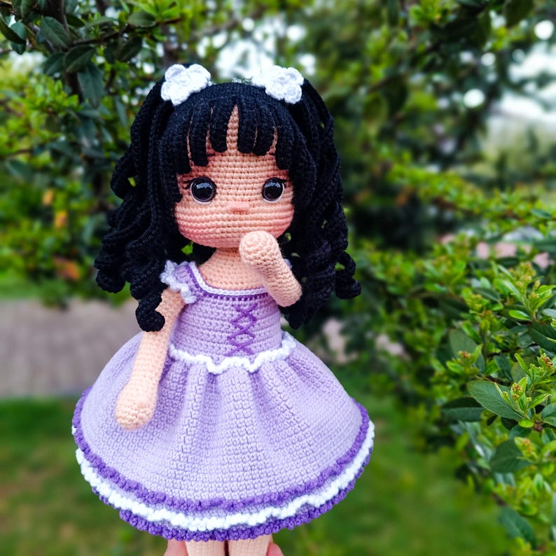K&ouml;nnte beinhalten: Eine geh&auml;kelte Puppe mit schwarzen Locken, die ein lila Kleid mit wei&szlig;en Verzierungen tr&auml;gt. Die Puppe hat gro&szlig;e braune Augen und einen s&uuml;&szlig;en Ausdruck.