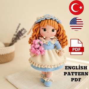 Pode incluir: Uma boneca de crochê feita à mão com cabelo encaracolado ruivo, vestida com um vestido azul e creme, segurando um buquê de rosas cor-de-rosa. A boneca tem um chapéu azul e sapatos a condizer. A imagem inclui o texto "ENGLISH PATTERN PDF".