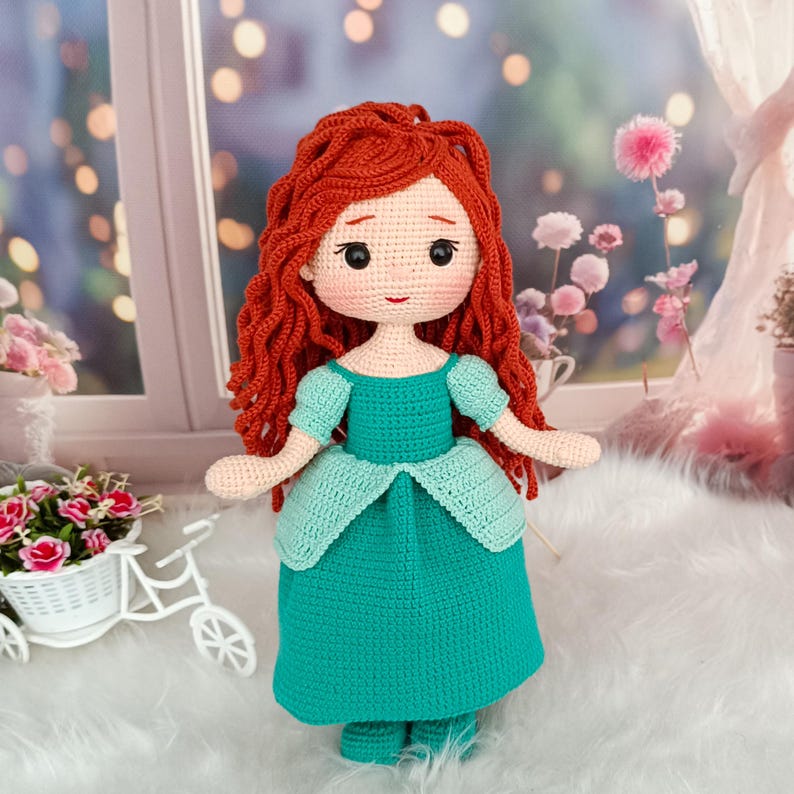 Op de afbeelding: Een handgemaakte gehaakte pop met rood haar en een zeegroene jurk. De pop heeft een lieve uitdrukking met roze wangen en zwarte ogen. De jurk heeft een gelaagde rok en korte mouwen. De pop is ongeveer 30 cm hoog.
