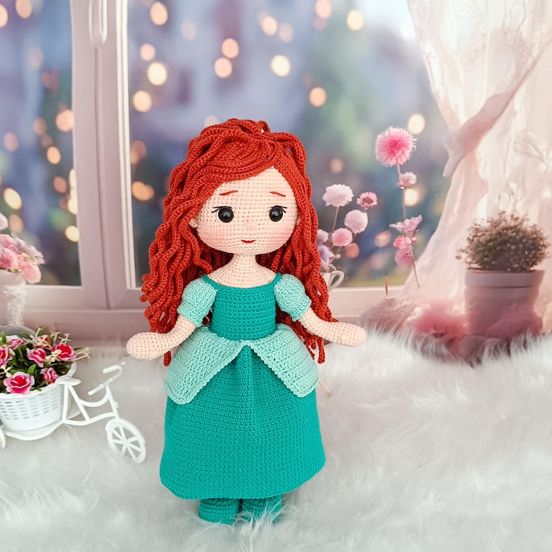 Op de afbeelding: Handgemaakte gehaakte pop met rood haar en een turquoise jurk. De pop heeft een lieve uitdrukking met zwarte ogen en een kleine glimlach. De jurk heeft een licht turquoise lijfje en een donkerdere turquoise rok. De pop staat voor een raam.