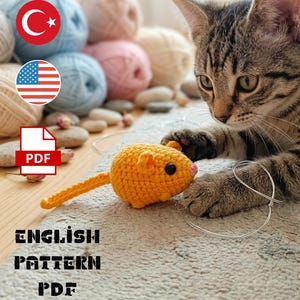 May include: An orange crocheted mouse toy with a long tail and black eyes sits on a rug. A tabby cat is focused on the toy. The image includes balls of yarn in various colors and the text "ENGLISH PATTERN PDF".