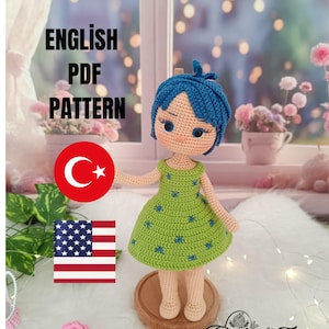 以下が含まれることがあります： 青い髪と青い星模様の緑色のドレスを着た手作りの編み人形。人形は木の台座の上に立っています。画像には「ENGLISH PDF PATTERN」というテキストとトルコと米国の国旗が含まれています。