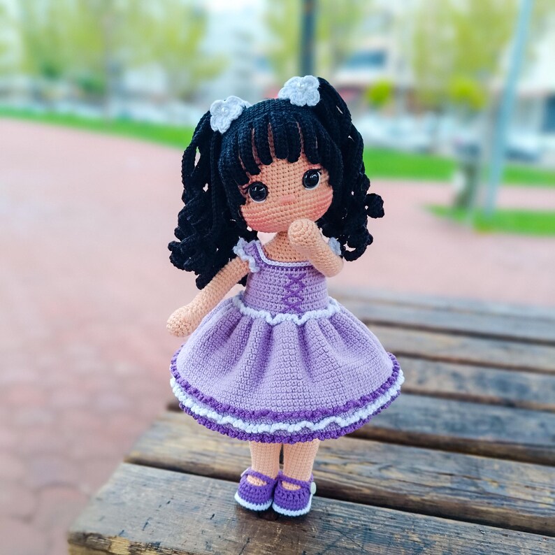 K&ouml;nnte beinhalten: Eine geh&auml;kelte Puppe mit schwarzen lockigen Haaren, wei&szlig;en Schleifen und einem lila Kleid mit wei&szlig;er Spitzenborte. Die Puppe tr&auml;gt lila Schuhe und steht auf einer Holzoberfl&auml;che.