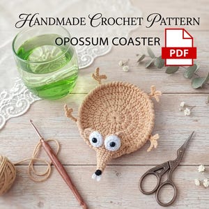May include: Handmade crochet pattern for an opossum coaster. The tan coaster has a circular design with a long snout, large eyes, and small feet. A glass of green liquid, crochet hook, scissors, and yarn are also in the image.