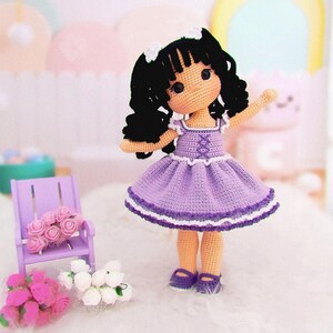 K&ouml;nnte beinhalten: Eine geh&auml;kelte Puppe mit langen, schwarzen, lockigen Haaren, die ein lila Kleid mit wei&szlig;er Spitzenborte und lila Schuhen tr&auml;gt. Die Puppe sitzt auf einer wei&szlig;en Oberfl&auml;che mit rosa und wei&szlig;en Blumen.