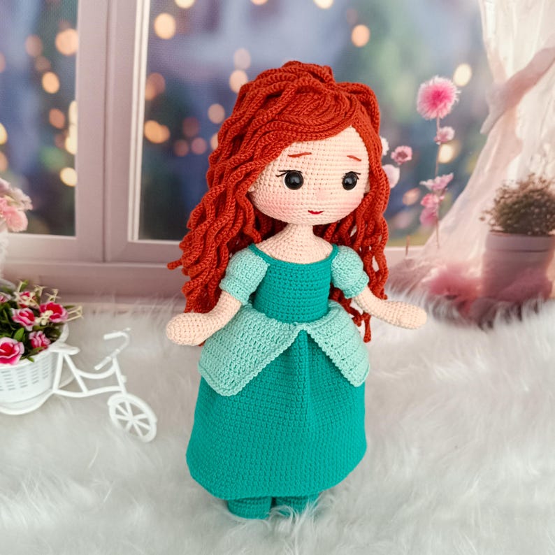 Op de afbeelding: Een handgemaakte gehaakte pop met rood haar en een teal jurk. De pop heeft een gedetailleerd gezicht met zwarte ogen en een kleine glimlach. De jurk heeft een licht teal lijfje en rokaccenten. De pop is ongeveer 30 cm hoog.
