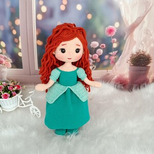 Op de afbeelding: Een handgemaakte gehaakte pop met lang, krullend rood haar en een turquoise jurk. De pop heeft een lieve uitdrukking met zwarte ogen en een kleine rode mond. De jurk heeft een geschulpte halslijn en mouwen. De pop staat op een wit oppervlak.