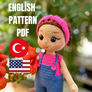 May include: A crocheted doll wearing a pink top and blue dungarees, with a pink headband. The doll has black eyes and brown hair. The image includes the text "ENGLISH PATTERN PDF" and flags of Turkey and the United States.