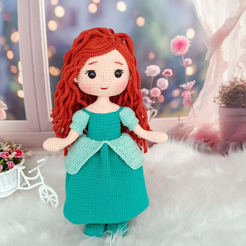 Op de afbeelding: Een handgemaakte gehaakte pop met rood haar en een turquoise jurk. De pop heeft een gedetailleerd gezicht met zwarte ogen en een kleine rode mond. De jurk heeft een geschulpte rand en korte mouwen. De pop staat voor een vage achtergrond.