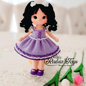 K&ouml;nnte beinhalten: Eine geh&auml;kelte Puppe mit schwarzen Locken, die ein lila Kleid mit wei&szlig;en Verzierungen und lila Schuhe tr&auml;gt. Die Puppe sitzt auf einer wei&szlig;en Spitzenoberfl&auml;che mit drei Tulpen im Vordergrund.