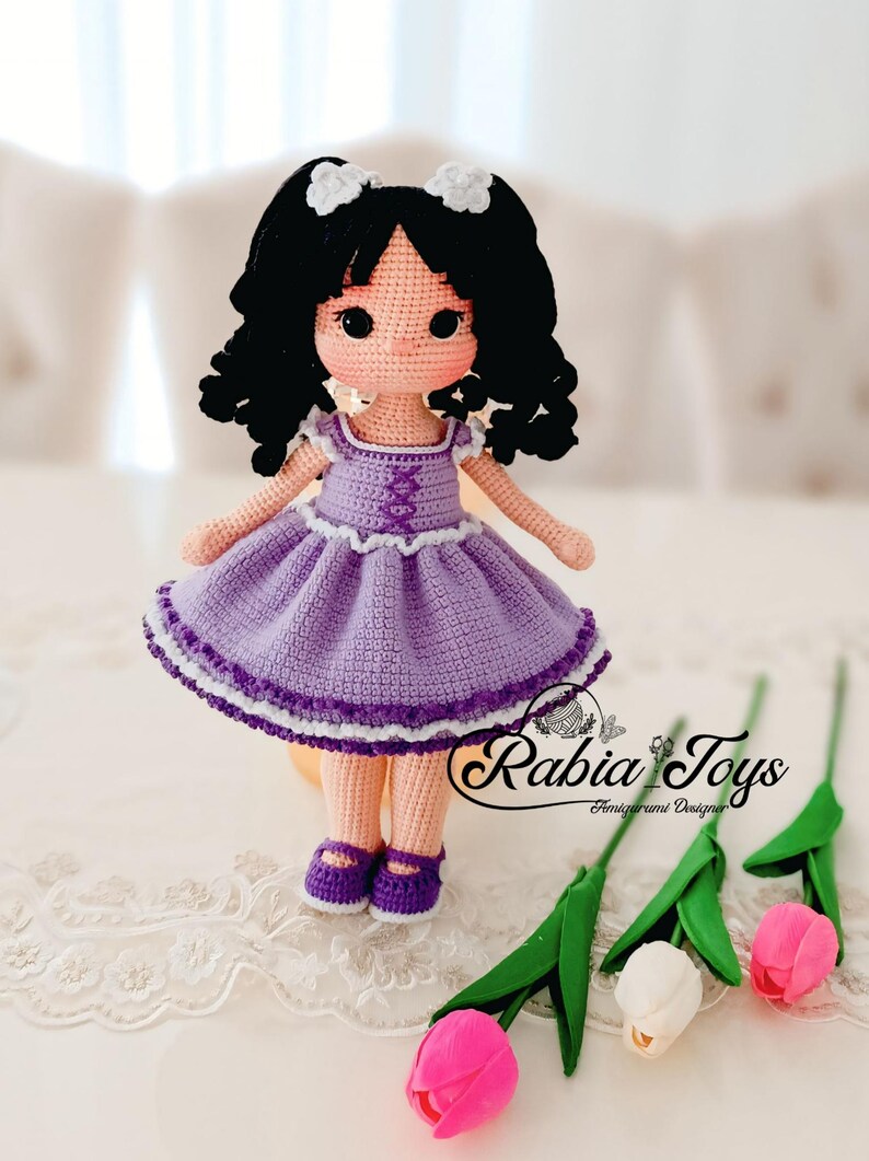 K&ouml;nnte beinhalten: Eine geh&auml;kelte Puppe mit schwarzen Locken, die ein lila Kleid mit wei&szlig;en Verzierungen und lila Schuhe tr&auml;gt. Die Puppe sitzt auf einer wei&szlig;en Spitzenoberfl&auml;che mit drei Tulpen im Vordergrund.