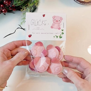 Pode incluir: Um saco de plástico transparente cheio de doces cor-de-rosa em forma de porco. Um cartão com a palavra alemã "Glücks-" e uma ilustração de um porco é visível. Os doces são segurados por uma pessoa.