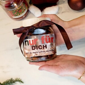Pode incluir: Um frasco de vidro cheio de uma substância escura, possivelmente um doce, é amarrado com uma fita marrom. O frasco tem uma tampa branca e um rótulo com as palavras "nur für DICH" em vermelho e branco. O frasco é segurado nas mãos.