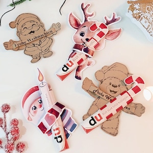 Peut inclure: Découpes sur le thème de Noël avec le Père Noël, un renne, une bougie et un ours en peluche, chacun tenant un sucre d'orge. Les découpes comportent du texte allemand, dont "Frohe Weihnachten" et "du bist voll". Décorations de vacances.