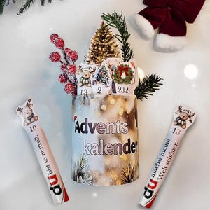 以下が含まれることがあります： 「Adventskalender」の文字が側面に書かれたアドベントカレンダー。ミニチュアのクリスマスツリー、トナカイ、番号付きのタグで飾られています。トナカイのイラストとドイツ語のテキストが入った2本のキャンディスティックが両側にあります。赤いリボンと緑の小枝がホリデーシーンを完成させています。