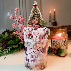 Peut inclure: Décoration de Noël cylindrique avec le texte allemand "Süße Weihnachtsüberraschung". Elle présente un sapin de Noël et des découpes de rennes, avec une bougie allumée et de la verdure festive en arrière-plan. La décoration évoque les fêtes de fin d'année.
