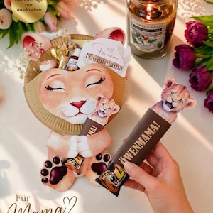 Könnte beinhalten: Ein papierner Löwen-Geschenkkorb mit einem lächelnden Gesicht, der einen Schokoriegel mit der Aufschrift "LÖWENMAMA!" hält. Der Korb ist mit kleinen Geschenken gefüllt und steht in der Nähe einer brennenden Yankee Candle und Blumen. Auf dem Korb steht "Für Mama".