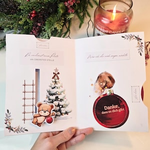 Può includere: Un biglietto di auguri a tema natalizio. Il lato sinistro presenta un albero di Natale e un orsacchiotto che tiene un ornamento rosso. Il lato destro ha un ornamento rosso con il testo "Danke, dass es dich gibt".