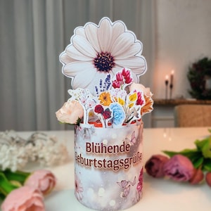 May include: A decorative floral arrangement featuring a large daisy and a variety of colorful paper flowers. The cylindrical container has the text "Blühende Geburtstagsgrüße" and butterfly accents. A perfect gift for a birthday.