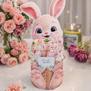 Puede incluir: Decoración de conejito de Pascua rosa con una cesta de conejitos y flores. El conejito tiene ojos grandes y una cara sonriente. La cesta tiene las palabras "Frohe Ostern" en una tarjeta pequeña. Rosas rosas y otras flores están en el fondo.