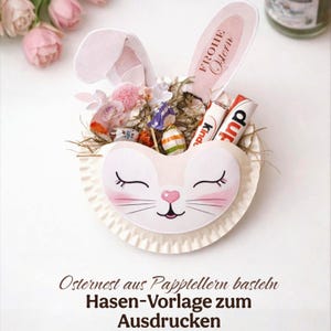 Puede incluir: Una cesta de Pascua decorativa con forma de cara de conejo, llena de caramelos, flores y dos barritas de chocolate. El conejo tiene mejillas rosas y ojos cerrados. El texto "FROHE Ostern" está en una de las orejas del conejo. El texto "Osternest aus Papptellern basteln Hasen-Vorlage zum Ausdrücken" está en la parte inferior.