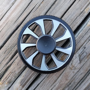 May include: A black and silver fidget spinner with a wheel design. The spinner has a black center and eight silver spokes.