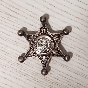 May include: A silver-colored sheriff badge in a star shape. The badge has a detailed design with the word "SHERIFF" in the center. The star points have small, round accents.