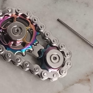 May include: A silver fidget spinner with two gears and a chain. The gears are iridescent and have a rainbow effect. The spinner comes with a small silver tool.