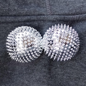 May include: Two silver massage balls with numerous small, pointed spikes. The balls are round and reflective, resting on a dark gray fabric surface. These massage tools are designed for muscle relief.