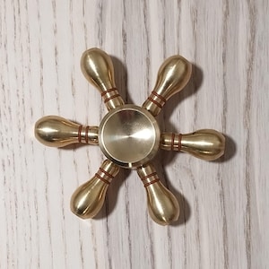 May include: A gold-colored metal fidget spinner with six arms. Each arm has a rounded end and a brown ring around the center.