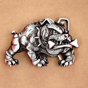 May include: A silver-colored metal belt buckle featuring a detailed bulldog design. The bulldog is depicted in a walking stance, holding a bone in its mouth. The buckle has a classic rectangular shape and a sturdy appearance.