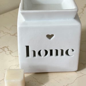 May include: A white ceramic wax melt warmer with a square top and a heart-shaped cutout. The word "home" is cut out below the heart. A small, square wax melt is in front of the warmer.