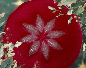 Peut inclure: Ornement de Noël rouge vif avec une fleur argentée scintillante au centre. L'ornement est entouré de feuilles vertes et de baies rouges, créant une ambiance festive. Le fond est vert doux avec des étoiles.