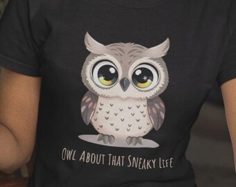 May include: A black short-sleeved t-shirt featuring a graphic of a wide-eyed owl with grey and white feathers. Below the owl, the text reads "OWL ABOUT THAT SNEAKY LIFE" in a simple, dark font. The shirt has a crew neckline and a standard fit.