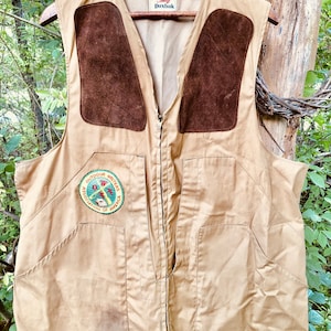 May include: Tan vest with brown suede shoulder patches and a full-length zipper. The vest has two front pockets and a patch that reads "Outdoor Writers Association of America". The brand name "Duxbak" is visible on the label.