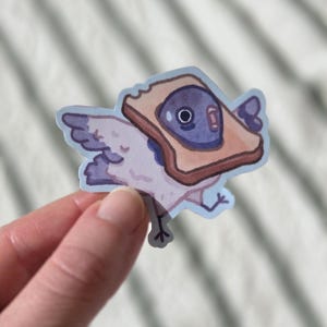 Puede incluir: Una pegatina caprichosa que representa un pájaro con una rebanada de pan tostado como cabeza. La tostada es marrón con un relleno de gelatina morada. El pájaro tiene alas moradas y un contorno azul claro.