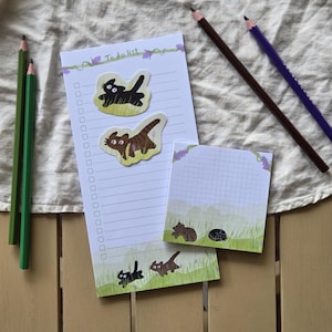 Peut inclure: Un bloc-notes rectangulaire ligné pour liste de tâches et un bloc-notes carré à carreaux, tous deux illustrés de chats dans de l'herbe verte. Deux crayons verts et deux crayons de couleur foncée sont disposés à côté de la papeterie.