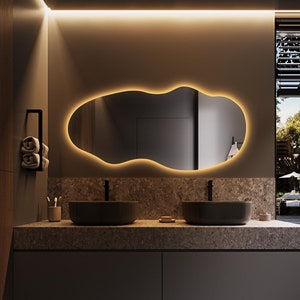 Può includere: Un mobile da bagno moderno con due lavabi e uno specchio grande di forma irregolare con illuminazione integrata. Il mobile è in legno scuro e ha un piano di lavoro di colore chiaro. Lo specchio è illuminato da una luce bianca calda.